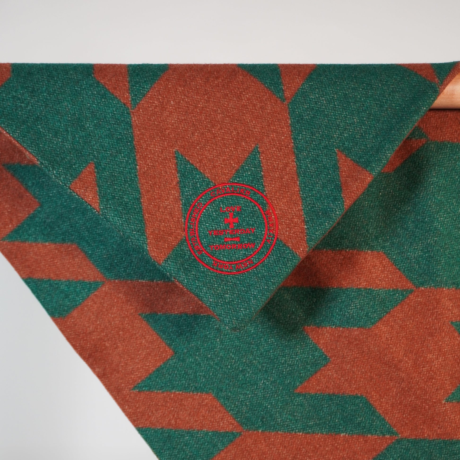 A close-up image of a corner of a wool blanket with a geometric pattern featuring a red embroidered emblem in the center.