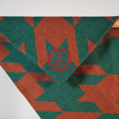 A close-up image of a corner of a wool blanket with a geometric pattern featuring a red embroidered emblem in the center.